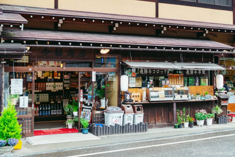 Traditional Japanese storefront with wooden architecture reminiscent of Shirakawa-go, shelves of bottles visible inside, food items and plants displayed outside, and lanterns hanging by the entrance on a rainy street.