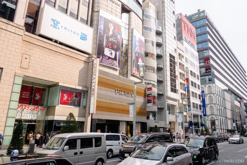 A busy street in Ginza, Tokyo, where shopping in Ginza thrives among modern buildings, shops, billboards, and parked cars. Pedestrians stroll the sidewalks as Japanese signs and advertisements fill the vibrant scene.