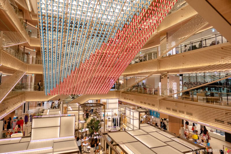 A modern shopping mall interior with spacious, well-lit walkways inspires the vibrant atmosphere of shopping in Ginza, featuring several floors, beige wooden accents, and red and blue hanging art suspended from the glass ceiling.