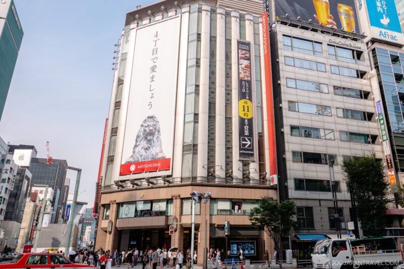 Busy street intersection in a city with tall buildings, including one with a large vertical banner featuring a dog and Japanese text. People enjoying shopping in Ginza walk on the sidewalks and cross the street under clear skies.