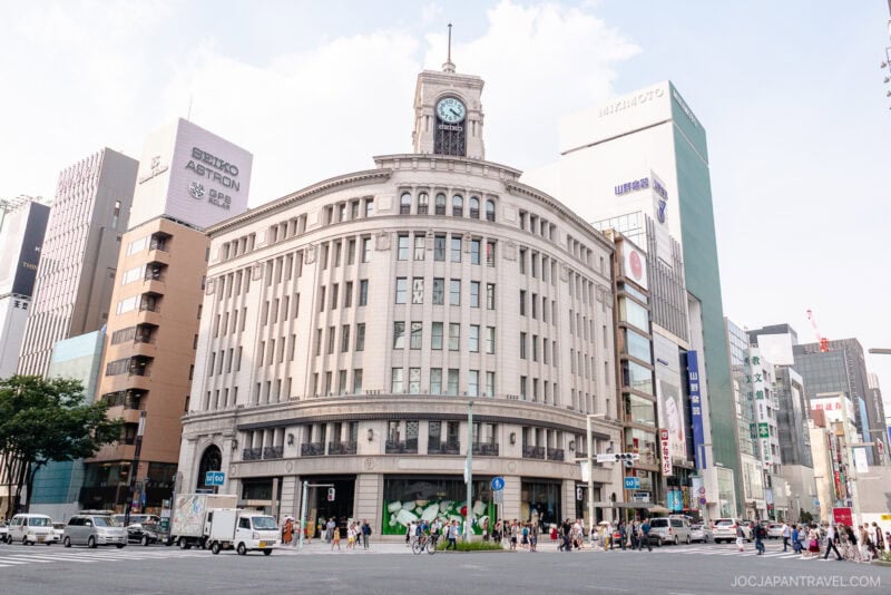 A large, historic building with a clock tower stands at a busy city intersection in Ginza, surrounded by modern tall buildings and people shopping in Ginza, walking across wide crosswalks in daylight.