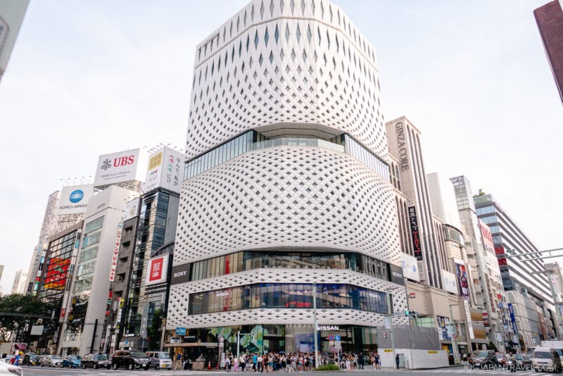 A modern, multi-story building with a unique, wavy white facade stands on a busy city street in Ginza, surrounded by commercial buildings and signs. People shopping in Ginza and cars are visible at street level.