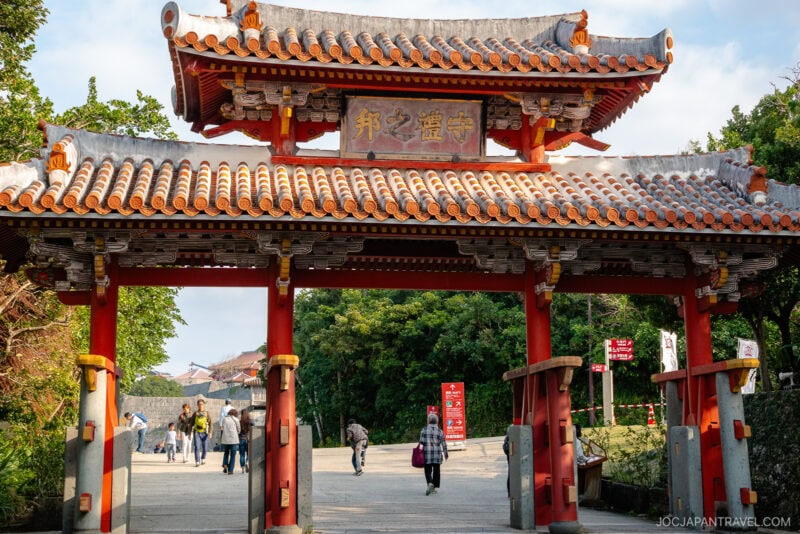 A traditional red and white Japanese gate with ornate roof tiles, reminiscent of Shuri Castle, stands at the entrance to a park; people are walking beneath it and trees are visible in the background.