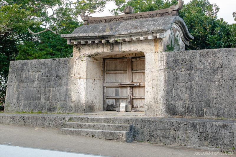 A traditional Japanese stone gate with a wooden door, reminiscent of Shuri Castle, stands between weathered stone walls, surrounded by green trees. Stone steps lead up to the closed gate.