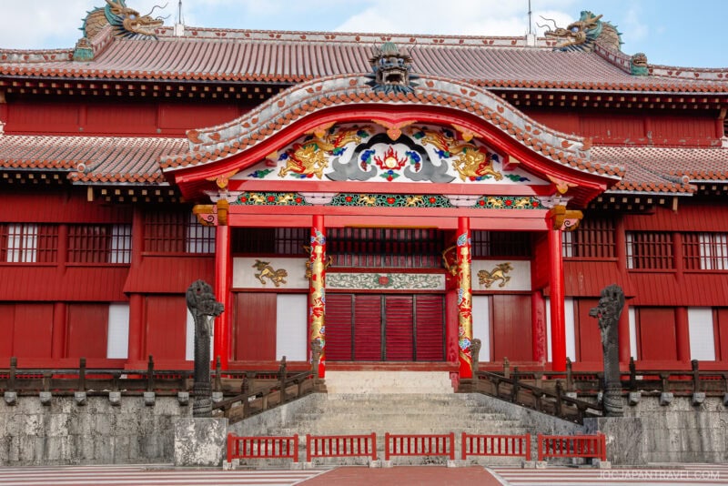 A traditional Japanese castle building inspired by Shuri Castle, featuring ornate red and gold decorations, intricate carvings, a curved roof, and wide stone steps leading to the entrance. The symmetrical structure is richly detailed.