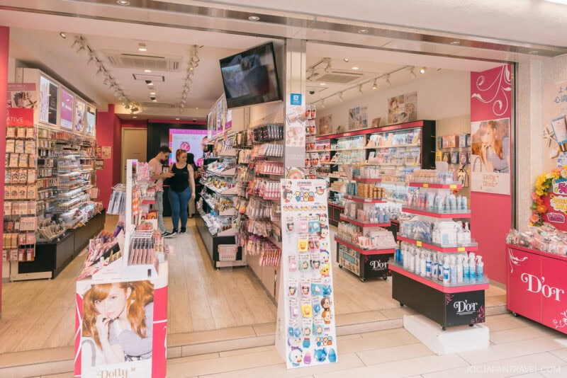 A brightly lit Japanese beauty store on Takeshita Street Harajuku, with shelves stocked with cosmetics, skincare, and beauty products. Two people browse inside. Pink and white decor create a cheerful atmosphere alongside promotional posters and display stands.