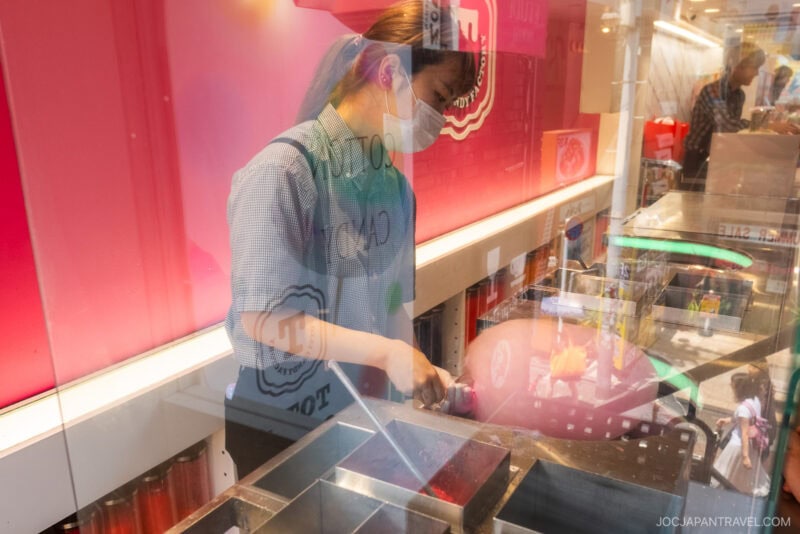A masked worker prepares food behind a glass counter inside a brightly lit shop on Takeshita Street Harajuku, with reflections of people and street activity visible on the glass.