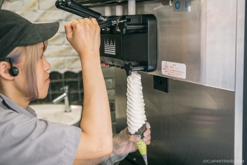 A worker in a gray uniform at Takeshita Street Harajuku dispenses a tall, spiral swirl of soft-serve ice cream into a cone from a machine, wearing a headset and gloves and focusing intently on making the ice cream.