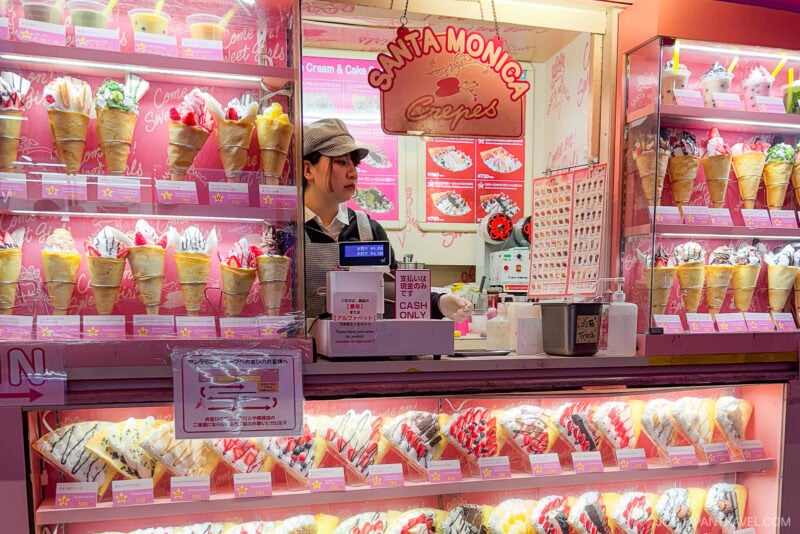 A crepe shop called Santa Monica Crepes on Takeshita Street Harajuku displays colorful crepes in plastic models. A worker stands behind the counter, ready to take orders. The shop has a bright, pink-themed interior.