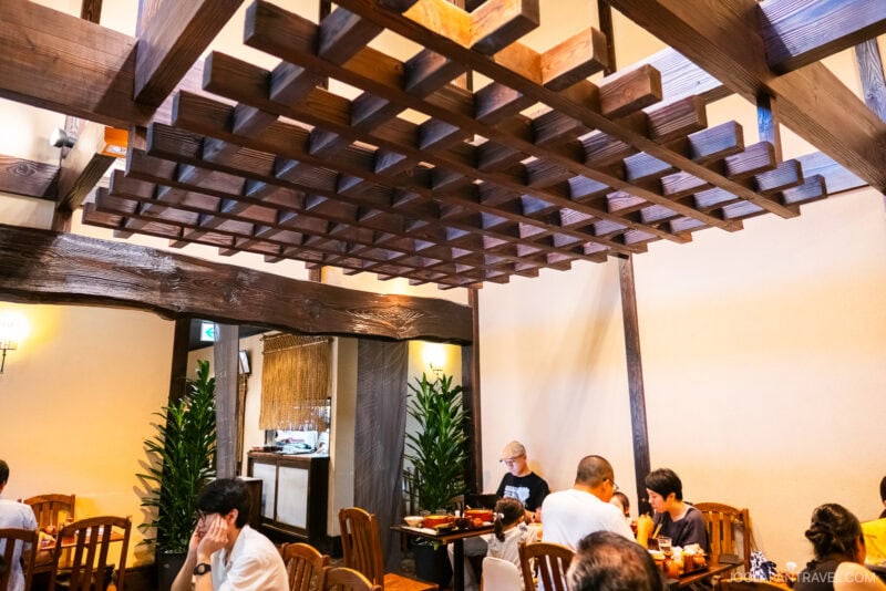 A cozy restaurant interior at Tamura Ginkatsu-tei features wooden beams and a lattice ceiling, wooden tables and chairs, warm lighting, and several people dining and conversing. Green plants decorate the walls for a welcoming touch.