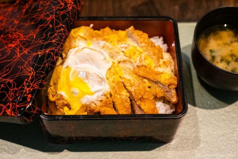 At Tamura Ginkatsu-tei, enjoy a lacquered bento box with breaded pork cutlet topped with egg over rice, partially covered by a decorative lid, and served with miso soup on a green placemat.