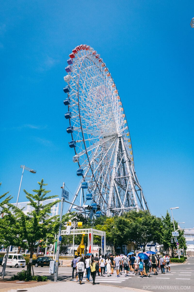 The Tempozan Giant Ferris Wheel rises against a bright blue sky, surrounded by green trees. People stroll along the street below, enjoying a sunny day at the amusement park.