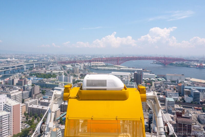 View from the top of a yellow Tempozan Giant Ferris Wheel gondola overlooking a cityscape with bridges, rivers, and distant buildings under a blue sky with scattered clouds.