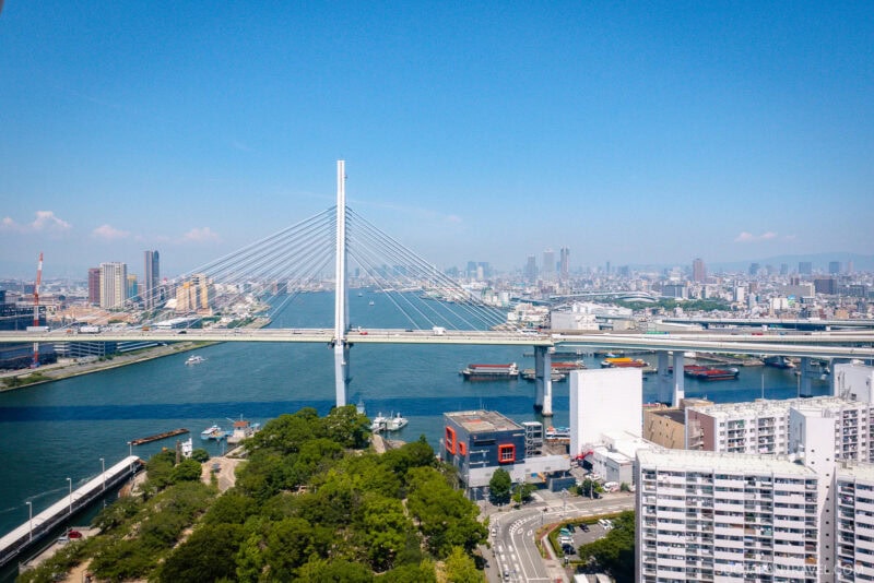 A modern cable-stayed bridge spans a wide river in a city, with boats on the water and tall buildings, including the Tempozan Giant Ferris Wheel, rising in the background under a clear blue sky. Green trees and various buildings line the riverbanks.