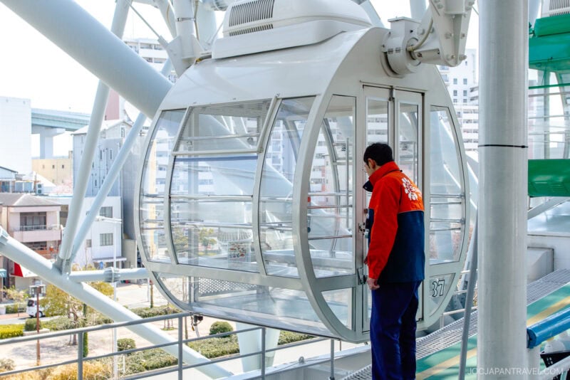 A person in a red and navy uniform stands beside a white Tempozan Giant Ferris Wheel cabin with glass windows, preparing to open the door. City buildings and greenery are visible in the background.
