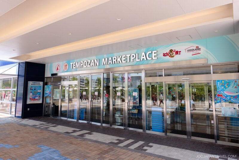 Entrance to Osaka Tempozan Marketplace with glass doors, signs for the food court, Legoland Discovery Center, and KidZania above. Light and dark tile patterns cover the floor, while reflections shimmer on the glass.