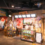 Tempozan Marketplace & Food Hall (天保山マーケットプレース) – Where Osaka’s Street Food and Retro Charm Come Alive A colorful Japanese restaurant entrance at Osaka Tempozan Marketplace features cherry blossom decorations, food displays, menu boards with Japanese text, and a life-sized statue of a man in a suit and hat amid bright lights and lively signage.