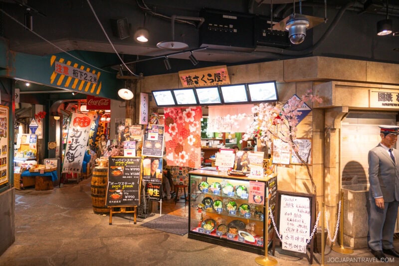 A colorful Japanese restaurant entrance at Osaka Tempozan Marketplace features cherry blossom decorations, food displays, menu boards with Japanese text, and a life-sized statue of a man in a suit and hat amid bright lights and lively signage.