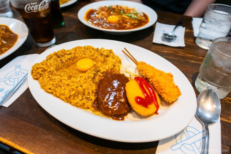 A plate of Japanese curry rice at Osaka Tempozan Marketplace is topped with a raw egg yolk, hamburger patty, fried shrimp, and croquette with ketchup, served alongside shredded cabbage. A glass of Coca-Cola, water, and utensils sit on the wooden table.