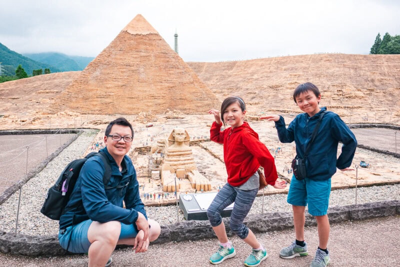 A man and two children pose playfully in front of miniature replicas of the Great Pyramid and Sphinx at Nikko Tobu World Square. The children mimic the Sphinx’s pose, smiling, while the man crouches nearby with a backpack.