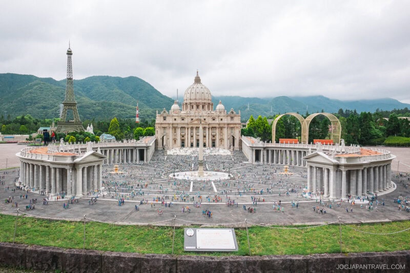 A detailed miniature model at Nikko Tobu World Square displays St. Peter’s Basilica and Square, with tiny crowds and nearby famous landmarks like the Eiffel Tower, all set against green mountains and a cloudy sky.