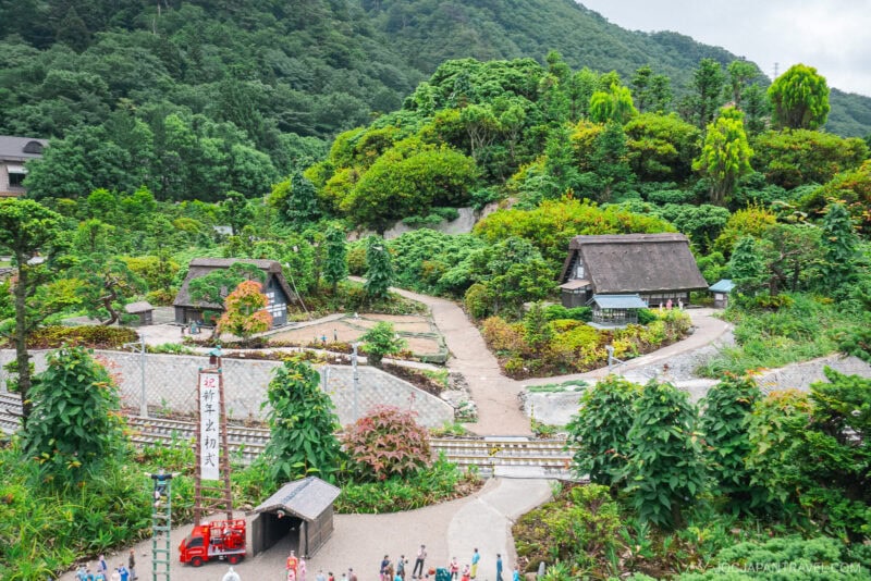A miniature village at Nikko Tobu World Square features winding paths, small traditional thatched-roof houses, lush green trees, and tiny figurines, all set against a backdrop of forested hills.