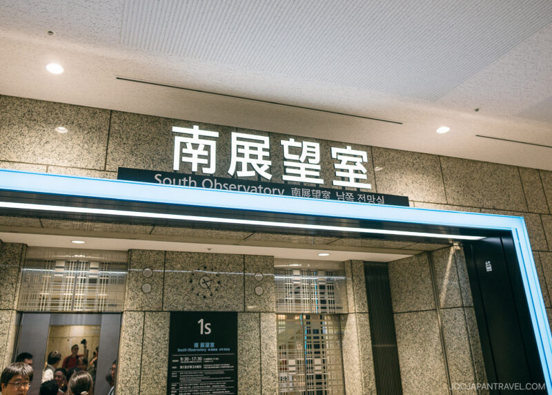 Entrance to the South Observatory at the Tokyo Metropolitan Government Building, with signs in English, Japanese, Chinese, and Korean above stainless steel doors. Several people enter the area, surrounded by beige tiled walls.