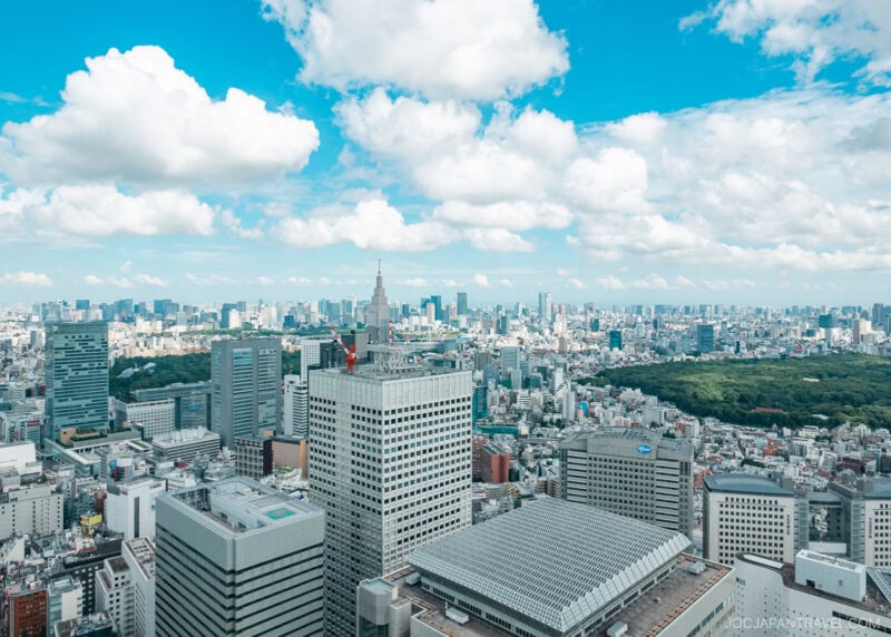 Aerial view of a modern cityscape featuring the Tokyo Metropolitan Government Building, tall skyscrapers, a lush green park, and scattered clouds in a bright blue sky.