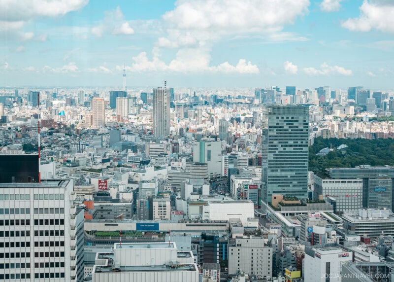 A wide view of a modern city skyline under a bright blue sky with scattered clouds, featuring numerous high-rise buildings, dense urban development, the Tokyo Metropolitan Government Building, and distant towers.
