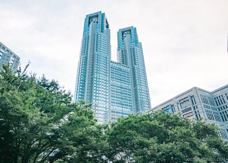 The image shows the iconic Tokyo Metropolitan Government Building, a tall modern skyscraper with twin towers, surrounded by green trees and adjacent buildings under a bright, partly cloudy sky.