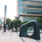 A modern city plaza in Tokyo Midtown with people walking, featuring a large abstract black sculpture with a hole in the center, trees, and sleek glass office buildings in the background.