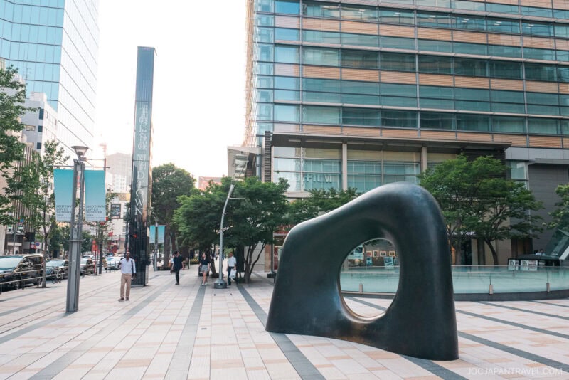 A modern city plaza in Tokyo Midtown with people walking, featuring a large abstract black sculpture with a hole in the center, trees, and sleek glass office buildings in the background.