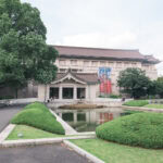 Exploring Tokyo National Museums at Ueno Park Tokyo National Museum stands behind a pond and manicured bushes, with a few people walking and sitting on benches nearby. The sky is partly cloudy.