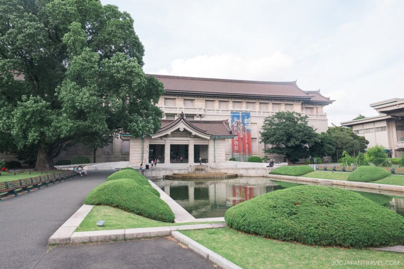 Tokyo National Museum stands behind a pond and manicured bushes, with a few people walking and sitting on benches nearby. The sky is partly cloudy.