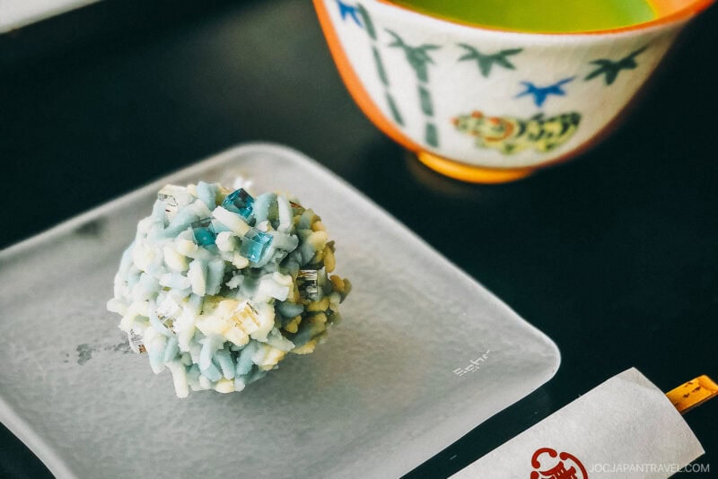 A close-up of a blue and white Japanese wagashi sweet from Toraya Tokyo on a glass plate, with a decorative cup of green tea in the background.