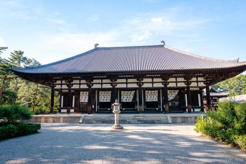 The traditional Japanese wooden temple building of Nara Toshodai-ji, with its large tiled roof, stands surrounded by greenery under a clear blue sky. A stone lantern is centered in front on the gravel courtyard.