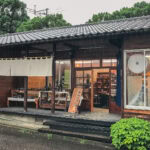 Touki Yufuin is a traditional Japanese pottery shop with a tiled roof, wooden exterior, large windows, and pottery displayed on a table outside. Its rainy, and lush greenery surrounds the building.