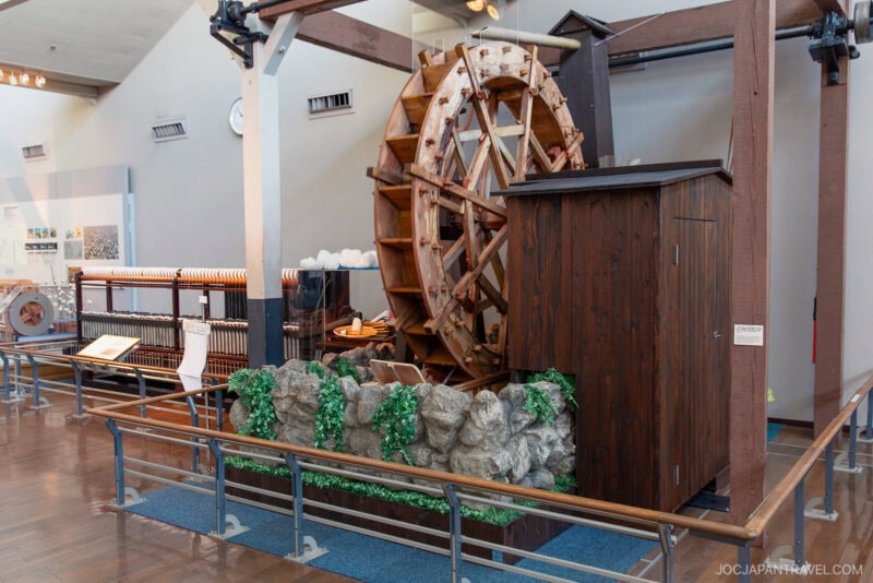 A large wooden water wheel is displayed indoors at the Toyota Commemorative Museum, surrounded by rocks and greenery, with an adjacent wooden structure. The well-lit exhibit features polished floors and informational displays in the background.