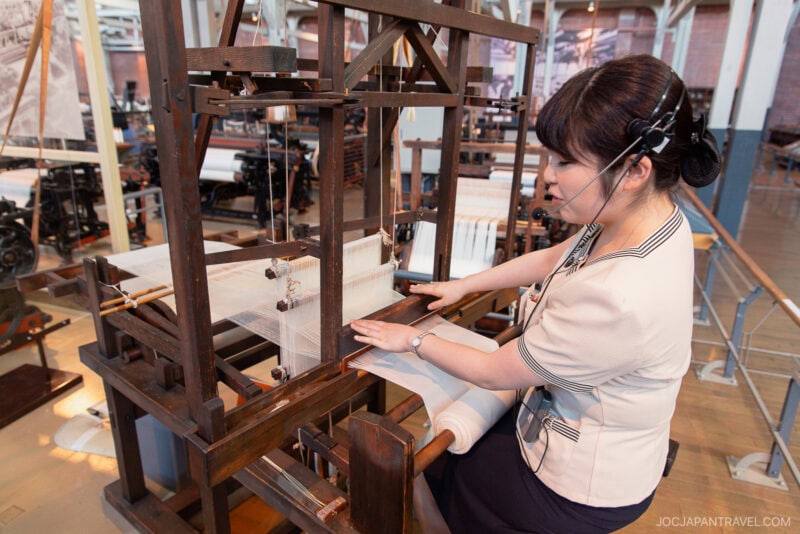 A woman wearing a headset demonstrates how to use a traditional wooden loom to weave fabric inside the Toyota Commemorative Museum exhibition space.