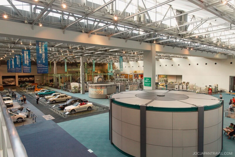 A spacious Toyota Commemorative Museum with vintage and modern vehicles on display, banners with Japanese text hanging from the ceiling, and a circular exhibit in the foreground under bright industrial lighting.