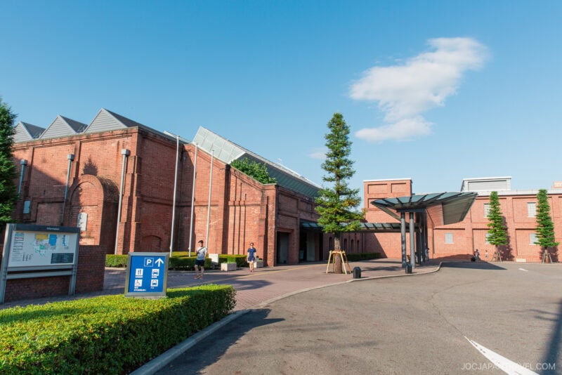 A large red-brick building with a modern canopy at the entrance, the Toyota Commemorative Museum welcomes visitors walking nearby, under a clear blue sky, with signage and greenery in the foreground.