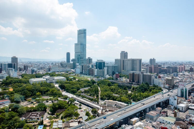 Aerial view of a modern cityscape featuring tall skyscrapers, lush green parks, and busy roads under a bright blue sky with scattered clouds, with the iconic Tsutenkaku Tower Osaka standing prominently in the skyline.