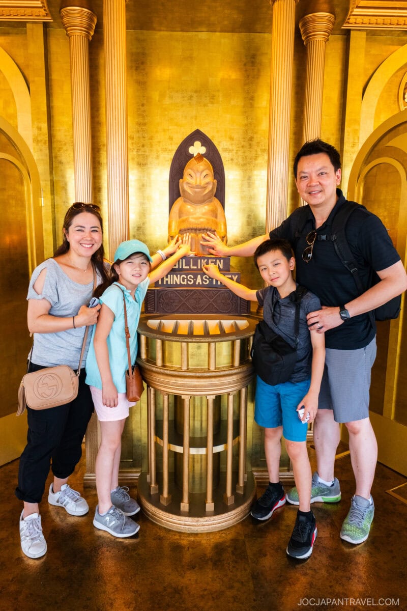 A smiling family of four poses in front of a golden Billiken statue inside a shrine at Tsutenkaku Tower Osaka, each touching the statue’s feet for good luck. The gold background glows as they all share a happy moment together.