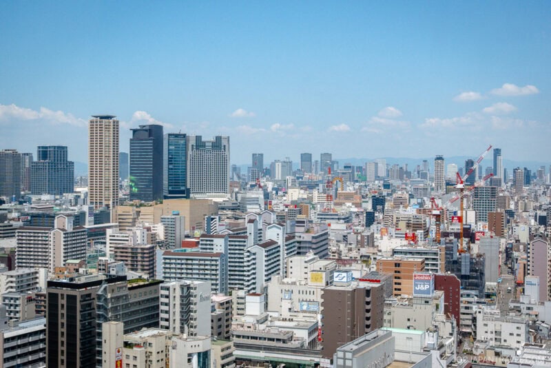 Aerial view of a modern cityscape with many tall buildings, skyscrapers, and construction cranes under a clear blue sky, featuring the iconic Tsutenkaku Tower Osaka in the skyline.