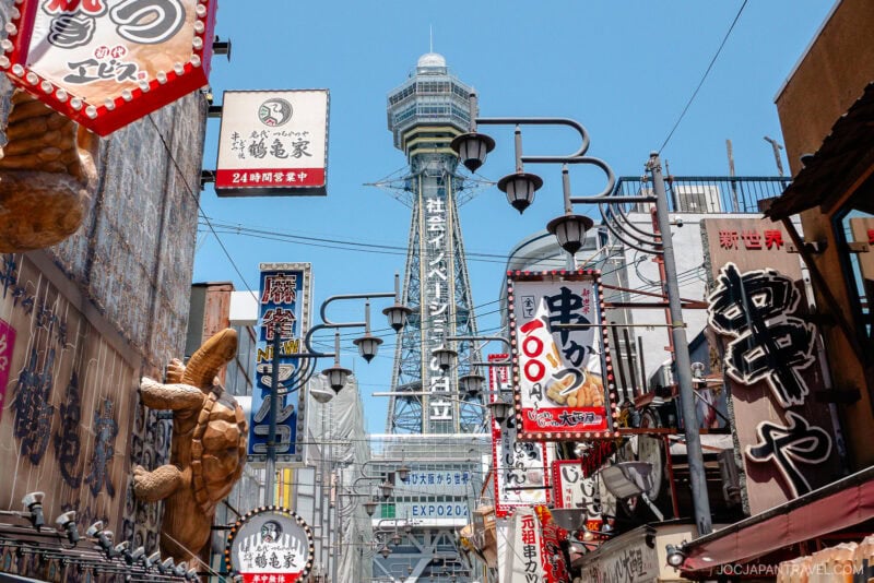 Colorful signs and lanterns line a bustling street in Osaka, with Tsutenkaku Tower Osaka rising in the background under a clear blue sky. Large food displays and Japanese text decorate this vibrant scene.