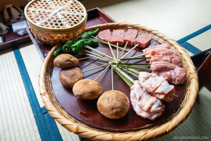 A woven tray on a tatami mat at Ukai Toriyama holds mushrooms, green peppers, sliced meat, and skewers, ready for grilling. A covered basket sits in the background.