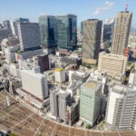 Umeda (梅田) – Shopping, Dining, and Travel at Osaka Station City Aerial view of Osaka Umeda’s bustling cityscape, showcasing tall modern skyscrapers, densely packed buildings, and crisscrossing railway tracks below under a bright blue sky with scattered clouds.