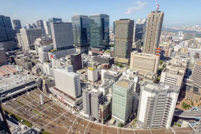 Aerial view of Osaka Umeda&rsquo;s bustling cityscape, showcasing tall modern skyscrapers, densely packed buildings, and crisscrossing railway tracks below under a bright blue sky with scattered clouds.