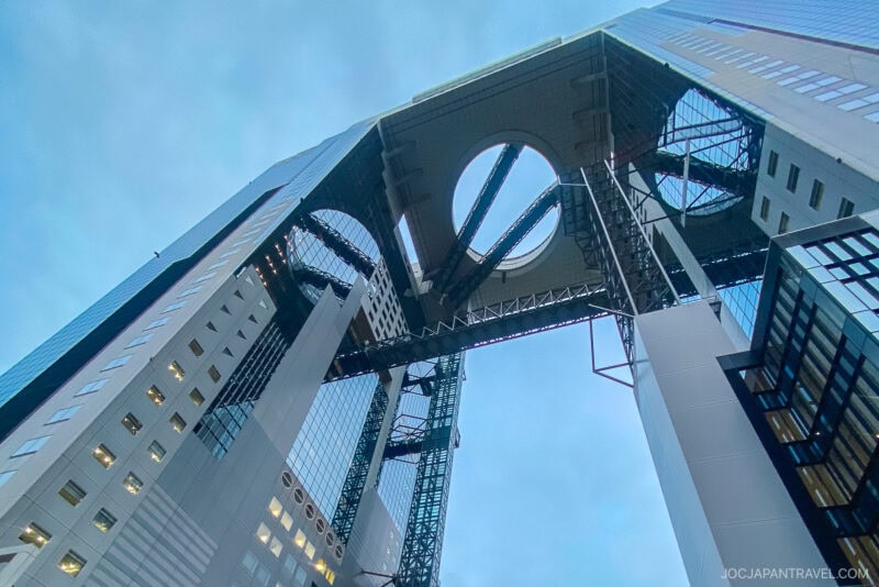 A dramatic upward view of the Umeda Sky Building in Osaka, Japan, showcases its two tall towers connected by a futuristic circular observation deck with escalators and bridges, all set against a cloudy sky.