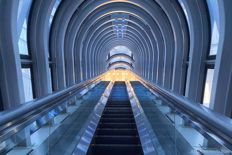 A futuristic glass-covered escalator ascends through an arched tunnel with curved metal beams and soft blue lighting, leading toward a bright, illuminated area at the top of the iconic Umeda Sky Building.